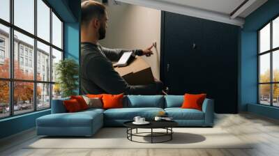 Back view of delivery man ringing doorbell of customer apartment holding cardboard box and contactless payment POS wireless terminal for card paying. Concept of online shopping. Wall mural