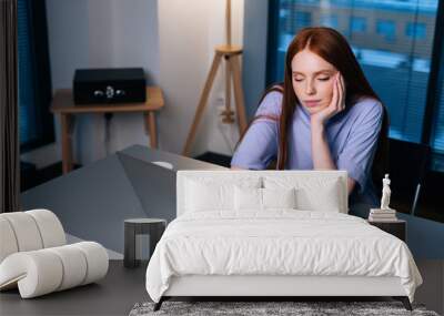 Bored young woman scrolling social networks at laptop sitting at desk in home office room near window. Tired redhead female working on computer at workplace evening at late. Wall mural