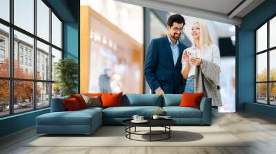 Cheerful beautiful young couple using mobile phone standing in hall of shopping mall. Bearded man in glasses and blonde woman buying at store centre. Concept of holiday sales, retail, consumerism. Wall mural