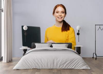 Cheerful young business woman or student holding laptop computer and looking at camera on isolated gray background. Pretty redhead lady model emotionally showing facial expressions, copy space. Wall mural