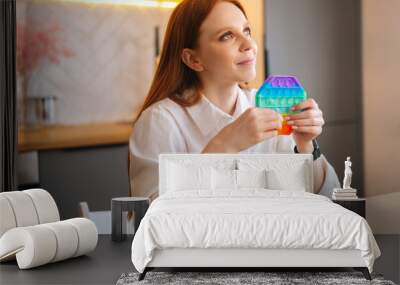 Close-up of dreaming charming redhead young woman pushing colorful iridescent soft silicone bubbles at home. Smiling female with trendy stress and anxiety relief fidgeting game in living room. Wall mural