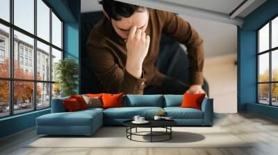 Close-up of stressed young man sitting alone feeling sad worry regret or fear, holding hand on forehead, depressed and desperate home. Suffering emotional pain and unhappy, mental health. Wall mural