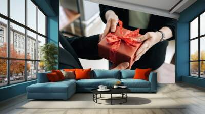 Close-up view of hands of unrecognizable woman giving red gift box tied to bow handed to man. Giving gifts during the Christmas, Happy New Year and Happy Birthday at office. Wall mural