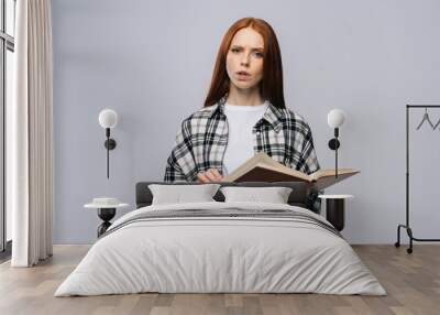 Concentrated serious young woman college student holding opened books and looking at camera on gray background. Pretty redhead lady model wearing casual clothes emotionally showing facial expressions Wall mural