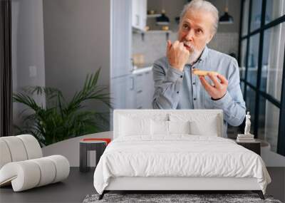 front view of handsome hungry bearded mature elderly male enjoying to eat pizza alone sitting at kitchen table. Front view of bearded senior older man with appetite to eat tasty pizza at home. Wall mural