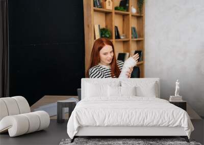 Front view of young woman with broken hand wrapped in white gypsum bandage having online video call, smiling looking to screen, working remotely on laptop computer sitting at desk in home office room Wall mural