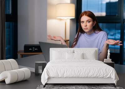 Frustrated young woman confused by laptop computer problem sitting at desk in home office near window evening at late, looking at camera. Bewildered female spreads hands incomprehensibly. Wall mural