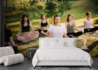 Group of mixed age women is practicing yoga and meditating morning in city park while sunrise. Group of people is sitting outdoors in lotus pose on grass with eyes closed Wall mural