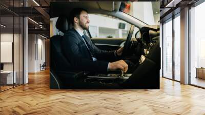 Happy bearded businessman wearing business suit is sitting behind wheel of new car in auto dealership. Concept of choosing and buying new car at showroom Wall mural