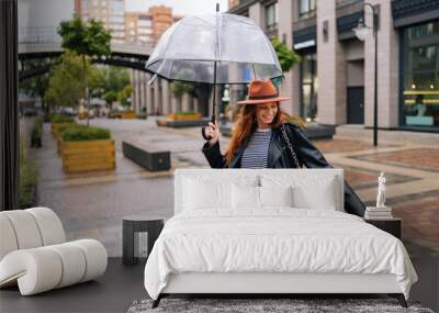 Happy excited young woman in elegant hat dancing and having fun with transparent umbrella on beautiful city street, enjoying rainy weather outdoors. Concept of female lifestyle at autumn season. Wall mural