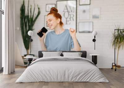 Happy redhead young businesswoman is using cell phone and laptop getting good while sitting at the desk in light cozy living room at home office. Wall mural