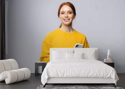 Happy smiling young woman holding mobile phone and looking at camera on isolated white background. Pretty redhead lady model emotionally showing facial expressions in studio, copy space. Wall mural