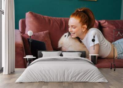 Happy young woman lying on comfortable sofa with white pretty spitz pet dog and using laptop in living room. Cute puppy licking nose of charming lady watching video from computer at home. Wall mural