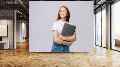 Happy young woman student holding laptop computer and looking away on isolated gray background. Pretty lady model with red hair emotionally showing facial expressions in studio, copy space. Wall mural