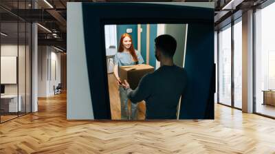 High-angle view of young woman receiving cardboard box from unrecognizable delivery man on doorstep at apartment. Back view of courier male delivering parcel package post to female client at home. Wall mural