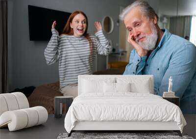 Joyful young woman and mature male having fun, enjoying leisure time, sharing news at home. Sad bearded grandfather talking with pretty young granddaughter sitting in bed. Wall mural