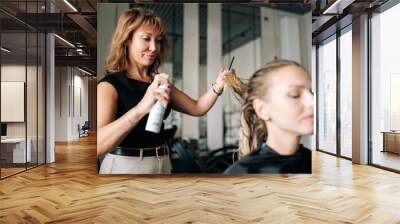 Lady client with long blonde hair getting stylish hairdo done at salon. Female hairdresser spraying hair setting liquid on blonde woman customer hair. Concept of professional fashion hairstyling. Wall mural