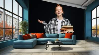 Laughing young woman college student holding books and looking at camera on isolated black background. Pretty redhead lady model wearing casual clothes emotionally showing facial expressions. Wall mural