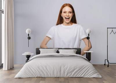 Laughing young woman wearing T-shirt and denim pants holds hands in pockets on isolated white background. Pretty lady model with red hair emotionally showing facial expressions in studio, copyspace. Wall mural