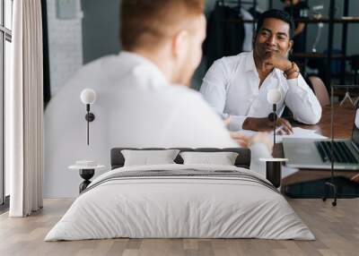 Pensive serious professional African American man interviewing caucasian European young candidate for job at successful company. Two business people talking strategy sitting at office desk. Wall mural