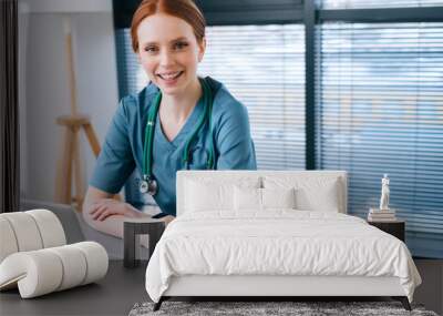 Portrait of attractive smiling young female doctor in blue green medical uniform sitting at desk with laptop on background of window in hospital office of medic clinic, looking at camera. Wall mural