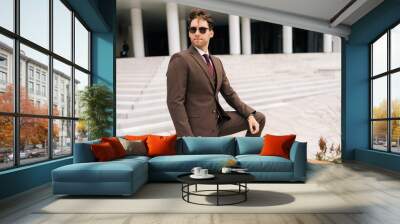 Portrait of confident successful businessman in brown suit and sunglasses standing in front of modern office building. Stylish businessman in elegant formal attire posing alone outdoors, looking away. Wall mural