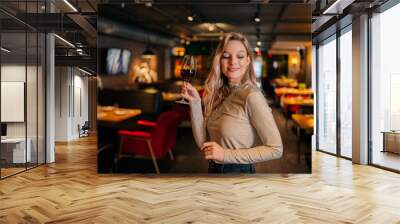 Portrait of elegant blonde woman holding glass of red wine standing in restaurant with luxury interior with dark interior, looking away. Front view of cute female posing at cafe, blurred background. Wall mural