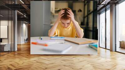 Portrait of exhausted pupil boy tired from studying holding head head with hands sitting at desk with paper copybook, looking down. Frustrated child schoolboy doing homework at home. Wall mural