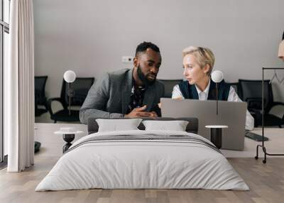 Portrait of focused multiethnic business partners looking at laptop screen, discussing corporate software application. Young black male intern employee consulting about project with female boss leader Wall mural