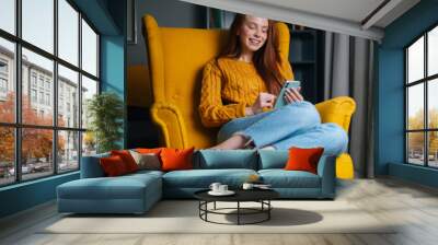 Portrait of happy redhead young woman using smartphone sitting in yellow armchair, smiling looking to device screen. Attractive ginger female holding phone in hands sitting in soft cozy chair at home. Wall mural