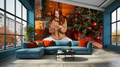 Portrait of happy young woman getting Christmas greeting card from her beloved on background of xmas tree at cozy living room with festive interior. Surprised lady pleasure reading letter at home. Wall mural