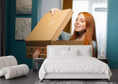 Portrait of happy young woman open box of hot pizza standing at doorway to apartment, looking inside. Hunger female customer received online order from courier with prepared meal for lunch Wall mural