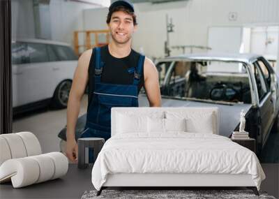 Portrait of positive mechanic male in uniform and cap standing posing auto service center, smiling looking at camera, enjoying time at workplace. Concept of professional automobile maintenance Wall mural