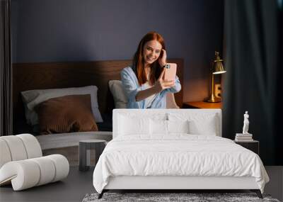 Pretty redhead young woman posing for mobile selfie in bed while sitting on bed at cozy bedroom. Caucasian female having video calling in mobile phone app at home. Wall mural