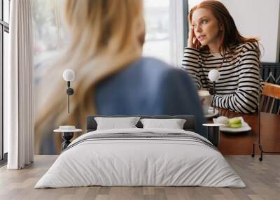 Rear view of unrecognizable blonde female talking to pensive woman friend, spending time together during drinking coffee at cozy cafe, sitting at table by window. Concept of woman friendship Wall mural