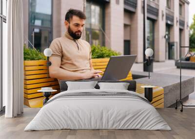 Serious bearded freelancer male working typing with laptop computer sitting on bench on city street with takeaway coffee. Confident young business man in casual typing on keyboard outdoors. Wall mural