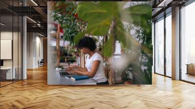 Side view of African young woman working typing on laptop computer sitting at desk in home office room with modern biophilia design, on background of green plants. Concept of house garden. Wall mural