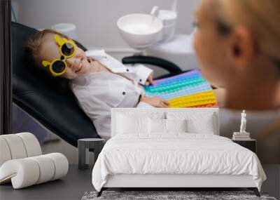 Side view of cheerful little child girl in funny eyeglasses playing with colorful pop it toy, sitting in dental chair during examining teeth from pediatric dentist. Concept of children teeth treatment Wall mural