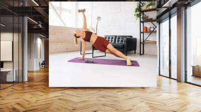 Strong fit young woman with perfect athletic body wearing sportswear doing side plank with dumbbell during workout training. Concept of healthy lifestyle and physical activity at home. Wall mural