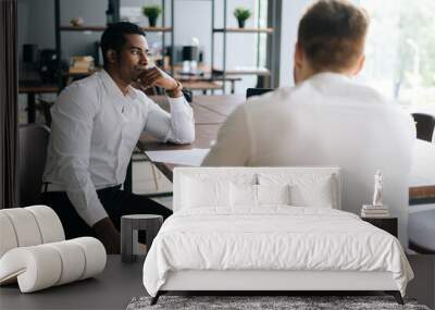 Thoughtful serious professional African American man interviewing caucasian European young candidate for job at successful company. Two business people talking strategy sitting at office desk. Wall mural