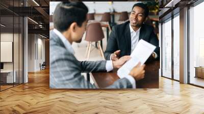 Two positive multiethnic coworkers sitting together at desk in modern office room and discussion over documents, close-up, selective focus. Concept of interracial friendship and cooperation. Wall mural