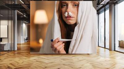 Vertical portrait of ill tired young woman suffering from runny nose disease with paper tissues into nose, measuring temperature with thermometer, sitting in bed covered with warm blanket at home. Wall mural