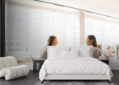 Wide shot of two female office workers sitting at desk with laptop looking at each other discuss project, brainstorming working together. Personnel manager interviewing job candidate in meeting room. Wall mural