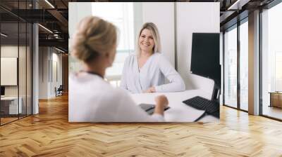 Young blonde woman at doctor reception in white dressing gown. Therapist female sitting at table and asks the patient questions Wall mural