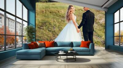 Full length body portrait of young bride and groom enjoying romantic moments outside at sunset in beautiful summer day. Wedding couple. Standing face to face with the green hills on background. Wall mural