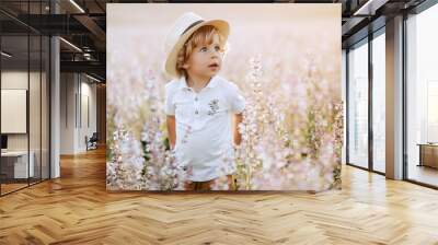 Happy little boy with blonde curly hair in pink sage field smiling, sunny day, outdoors. Child running on meadow in summer in nature. Kid with straw hat playing in wild flowers field at sunset. Wall mural