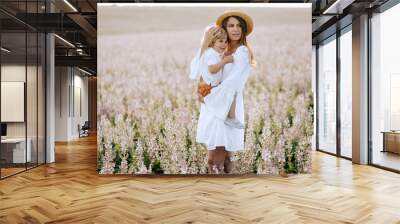 Mother and son playing in the field. Beautiful young woman wearing a white dress and straw hat playing with her little boy outside. Wall mural