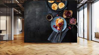 Pancakes with raspberry jam on a blue plate, frozen raspberries, a jar of raspberry jam, a cup of coffee, a frying pan with pancakes, fork and knife on dark metal table top view. Breakfast. Wall mural