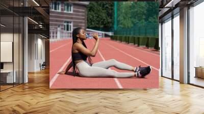 African American sportswoman sits on red running track and drinks bottled water in sunny morning. Black female runner rests after training and wants to show perfect results. Woman enjoys healthy life Wall mural