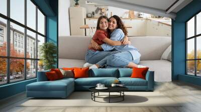 Carefree happy family of millennial woman and teenage girl hugging sits on big sofa in living room. Positive cheerful daughter hugging mother after long separation or arrival from school camp Wall mural